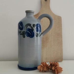 May include: A light grey ceramic jug with a single handle and a narrow neck. The body of the jug features a hand-painted floral pattern in shades of dark blue and green. The base of the jug is accented with a solid dark blue band around the bottom.