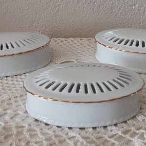 Puede incluir: Tres cajas ovaladas de porcelana blanca con tapa. Cada tapa presenta una serie de ranuras radiantes y un borde fino pintado en oro. La base de cada recipiente es lisa y blanca, combinando con el diseño de la tapa en un conjunto de tres.