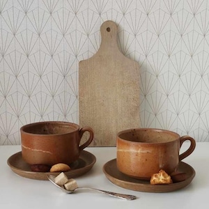 Peut inclure: Deux tasses à café en céramique marron avec soucoupes assorties, chacune contenant un biscuit et un chocolat. Une planche à découper en bois et une cuillère avec des cubes de sucre sont également présentes. Le fond présente un papier peint à motifs.