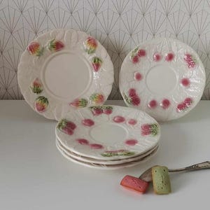 Könnte beinhalten: Ein Set aus fünf cremefarbenen Keramiktellern mit geprägten Fruchtmotiven, darunter Erdbeeren und Kirschen mit grünen Blättern. Die Teller haben einen erhöhten Rand und eine glatte Mitte. Ein silberner Löffel und zwei kleine rechteckige Kuchen sind enthalten.