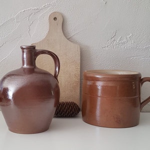 Könnte beinhalten: Eine braune Keramikkrug mit Henkel und eine braune Keramikbecher mit Henkel werden gezeigt. Ein Holzschneidebrett und ein Tannenzapfen befinden sich im Hintergrund. Der Krug ist rund mit schmalem Hals. Der Becher ist zylindrisch.