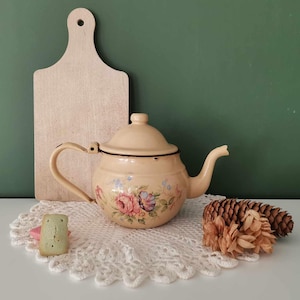 Puede incluir: Tetera de esmalte color crema con diseño floral de rosas, ribete negro y asa curvada. Una tabla de cortar de madera y un tapete de encaje blanco están en el fondo. También hay piñas decorativas y un pequeño objeto verde.