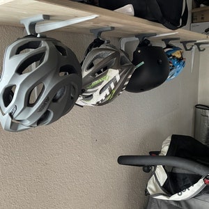 Puede incluir: Tres cascos de bicicleta colgados de una estantería blanca con ganchos grises. Los cascos son grises, negros y blancos con detalles verdes y azules.