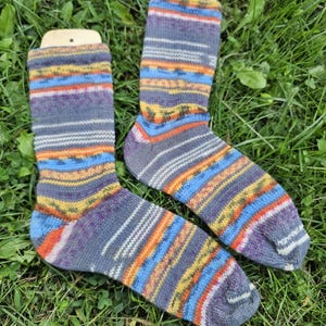 May include: A pair of colorful knitted socks with a striped pattern in shades of blue, orange, yellow, and purple. The socks are displayed on wooden sock forms and rest on a bed of green grass.