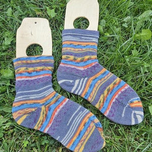 May include: A pair of hand-knitted socks displayed on wooden sock forms. The socks feature a striped pattern in shades of blue, purple, orange, yellow, and white. The socks are resting on a bed of green grass.