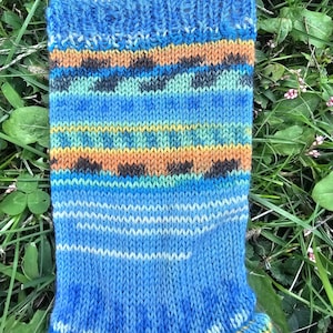 May include: A blue and multi-colored knitted sock with a wooden sock form. The sock features bands of orange, teal, and black geometric patterns. The cuff and toe are a solid blue color. The sock is displayed on a wooden form, set against a background of green grass.