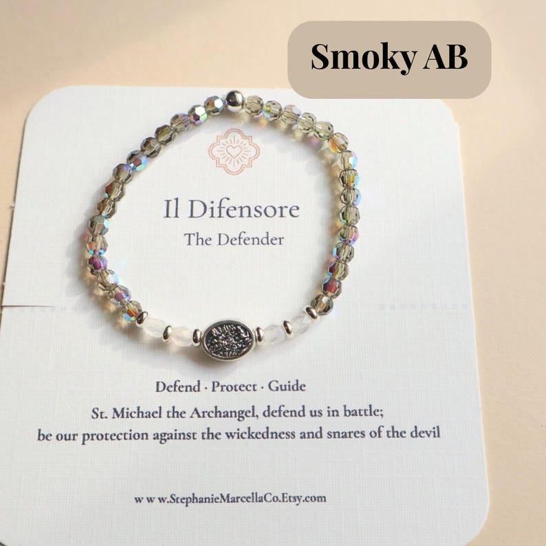 May include: Smoky AB beaded bracelet with the text "Il Difensore The Defender" on a white card. The bracelet features iridescent beads and a central decorative element. The card also includes the text "Defend Protect Guide".