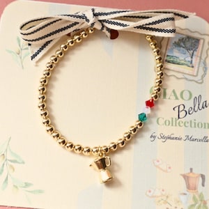 May include: Gold beaded bracelet with a charm of a coffee maker. The bracelet has a small section of red, white, and green beads, and is tied with a striped bow. The text "CIAO Bella Collection" is visible on the background.