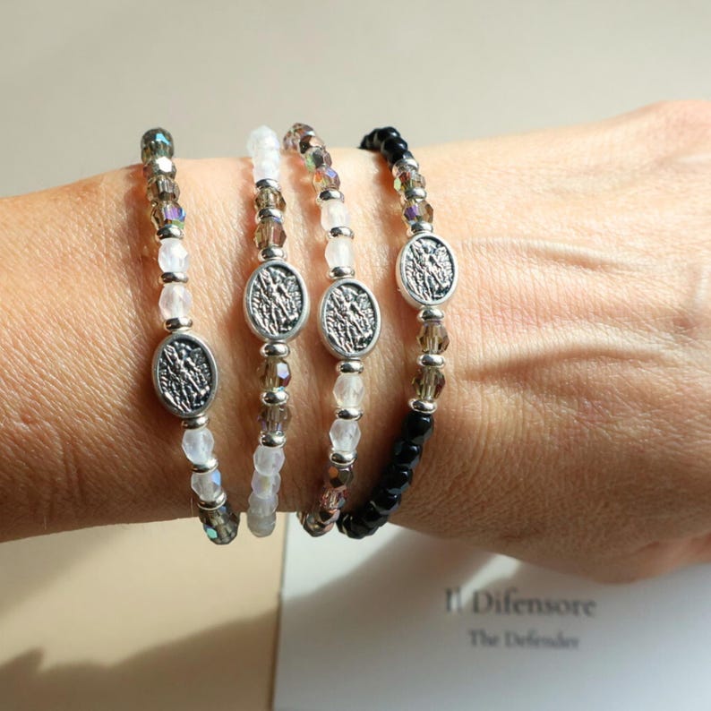 May include: Four beaded bracelets with silver oval charms are displayed. The bracelets feature a mix of clear, brown, and black beads. Each bracelet has a silver oval charm with a detailed design.
