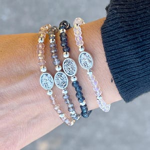 May include: Four beaded bracelets with silver angel charms. The bracelets are layered on a wrist and feature different colored beads: light pink, light brown, dark gray, and black.