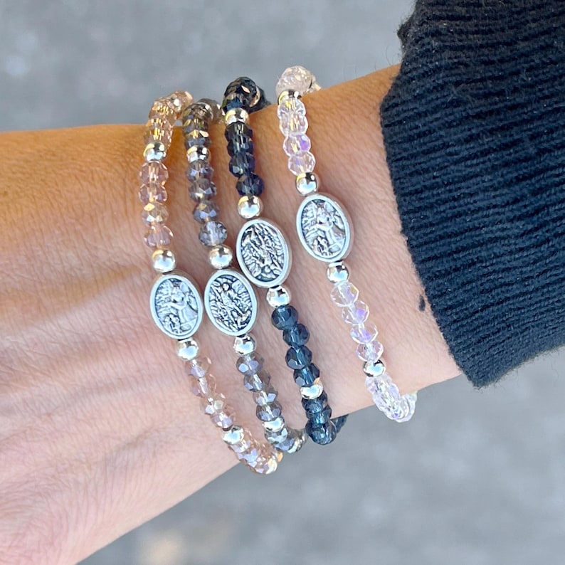 May include: Four beaded bracelets with silver angel charms. The bracelets are layered on a wrist and feature different colored beads: light pink, light brown, dark gray, and black.
