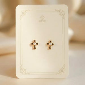May include: A pair of gold-colored cross stud earrings with a small white pearl in the center of each cross. The earrings are displayed on a cream-colored card with decorative borders and the text "STEPHANIE MARCELLA".