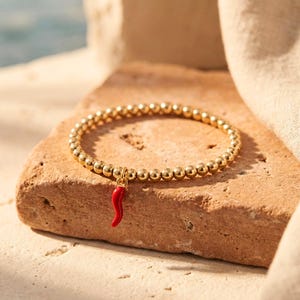 May include: A gold-toned beaded bracelet featuring a small, curved red chili pepper charm. The bracelet is constructed from a series of uniform, polished metallic spheres strung together to form a circular band with a single pendant attached.