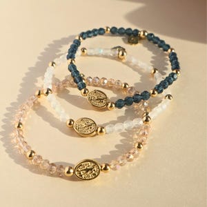 May include: Three beaded bracelets in shades of pink, white, and navy blue. Each bracelet features small, round beads and gold-colored accents. The bracelets have a central gold-colored oval charm with a detailed design.