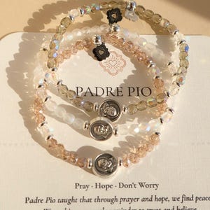 May include: Three beaded bracelets with iridescent and neutral-toned beads. Each bracelet features a silver charm with a religious figure and a small black flower charm. The text "PADRE PIO" is visible on the card.