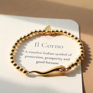 May include: Gold-colored beaded bracelet featuring an Italian horn charm. The charm is a stylized cornicello, a traditional Italian symbol. The card behind the bracelet reads "Il Corno" and describes it as a symbol of protection, prosperity, and good fortune.
