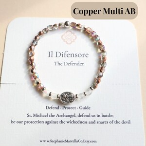 May include: A beaded bracelet with copper and iridescent crystals. The bracelet features a silver-toned oval charm with an engraved design. The text "Il Difensore The Defender" is visible on the card. The bracelet is displayed on a white card with additional text.