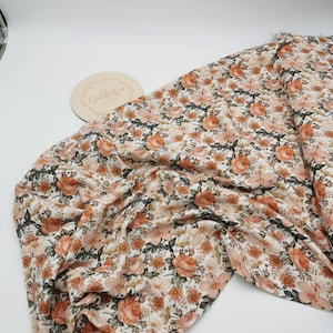 Könnte beinhalten: Ein floraler Stoff mit cremefarbenem Hintergrund und einem Muster aus pfirsichfarbenen und weißen Rosen, Gänseblümchen und anderen Blumen. Der Stoff ist über eine weiße Oberfläche drapiert. Eine Holzscheibe mit dem Text "Goldleafco" ist sichtbar.