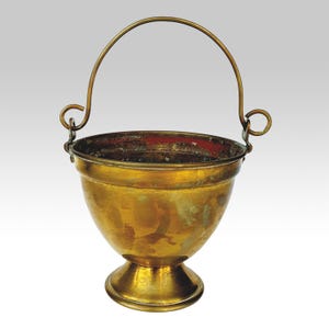 Op de afbeelding: Een vintage messing emmer met een gebogen handvat. De emmer heeft een afgeronde body die taps toeloopt naar een voetstuk. De binnenrand is rood. De handgreep is bevestigd met decoratieve gekrulde accenten.