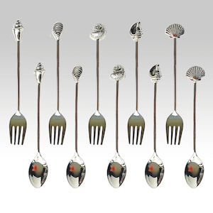 Puede incluir: Un conjunto de utensilios de servir plateados. Cada utensilio presenta un tenedor en un extremo y una cuchara en el otro, con diseños decorativos de conchas marinas. Los diseños incluyen varias formas de conchas, añadiendo un tema costero.