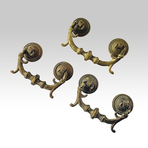 Può includere: Quattro maniglie antiche per cassetti in ottone con un design decorativo. Ogni maniglia presenta una piastra posteriore rotonda e una maniglia curva con dettagli ornati. Le maniglie sono di un caldo colore ottone invecchiato, che suggerisce uno stile vintage o retrò.