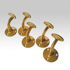 May include: Five brass handrail brackets. Each bracket has a circular base with three screw holes, a curved support, and a flat, oval top. The brackets are a warm, golden colour.