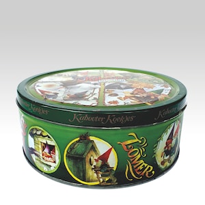 May include: A circular green tin with a lid, decorated with illustrations of gnomes and woodland scenes. The tin is labelled with the words "Kabouter Koekjes" and "Lomer". The tin is designed to hold biscuits or other treats.