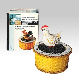Puede incluir: Un temporizador de cocina blanco y amarillo con forma de gallina, posado sobre una cesta de mimbre amarilla. El temporizador tiene un centro negro con números blancos y una cresta roja. La caja de al lado tiene texto en varios idiomas.