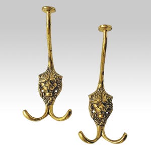 Può includere: Due ganci da parete in metallo color oro, ciascuno caratterizzato da un dettagliato design a testa di leone al centro. I ganci presentano un lungo e sottile collo verticale sormontato da una piastra di montaggio circolare e due bracci curvi.