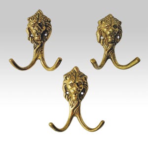May include: Three wall hooks made of antique brass-toned metal, each featuring a sculpted lion head at the top and two curved arms extending downwards to form a double hook design, with visible screw holes on the sides of each lion head.