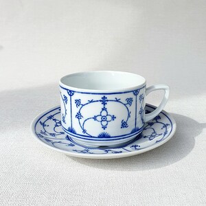 May include: White teacup and saucer set with a blue floral pattern. The cup has a handle and sits on a matching saucer. The design features stylized flowers and geometric shapes.