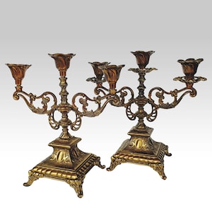 May include: Two antique brass candelabras with ornate floral designs. Each candelabra has multiple arms for candles, with a square base and decorative detailing. The candelabras are a warm gold color.