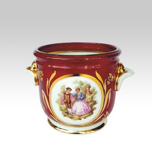 Pode incluir: Um recipiente de cerâmica vermelho profundo com duas alças laterais, apresentando um medalhão oval central que retrata duas figuras em trajes de época dentro de uma paisagem. O exterior é decorado com detalhes em ouro, motivos florais e borda dourada.