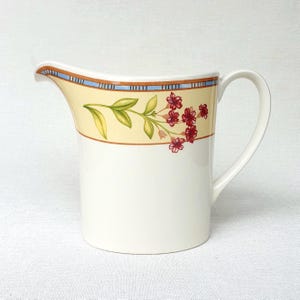 May include: A cream-coloured ceramic creamer with a floral design. The creamer has a yellow band with red flowers and green leaves, and a blue and brown border. It has a curved handle and a spout for pouring.