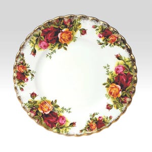 May include: A round white ceramic plate with a scalloped edge trimmed in gold. The plate features a floral pattern of red, pink, and yellow roses with green leaves arranged in clusters around the perimeter, leaving the center white.