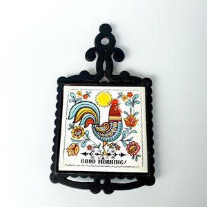 Könnte beinhalten: Ein schwarzer Metall-Topfuntersetzer mit einer dekorativen Kachel mit einem Hahn. Die Kachel zeigt einen bunten Hahn, Blumen und eine Sonne mit den Worten "GOOD MORNING!". Der Topfuntersetzer dient zum Schutz von Oberflächen vor heißem Kochgeschirr.