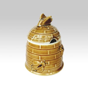 Può includere: Un vasetto di miele in ceramica marrone a forma di alveare, completo di coperchio e un piccolo foro per il cucchiaio. Il vasetto presenta un motivo a mattoni strutturato e decorazioni a forma di ape. Il coperchio è sormontato da una maniglia a forma di ape.