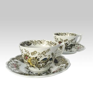 Könnte beinhalten: Zwei weiße Teetassen und Untertassen mit Blumen- und Vogelmotiven. Die Tassen und Untertassen haben einen gewellten Rand und sind mit gelben, braunen und blauen Akzenten verziert. Die Teetassen haben Henkel.