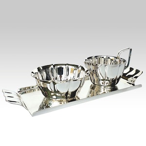 Pode incluir: Um conjunto de servir prateado com uma bandeja, uma tigela e uma chávena com pega. A tigela e a chávena têm um design de borda ondulada. A bandeja tem um design decorativo em forma de asa nas laterais. O conjunto é reflexivo e parece ser de metal.