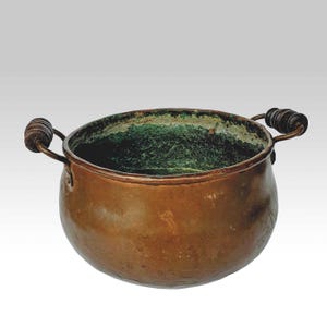 Könnte beinhalten: Ein Kupferkessel mit zwei gebogenen Griffen und Holzgriffen. Der Innenraum weist eine grüne Patina auf, während die Außenseite eine warme, gealterte Kupferoberfläche aufweist. Ideal für rustikale oder Vintage-Küchen.