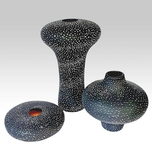 Op de afbeelding: Drie zwarte keramische vazen met een gespikkeld wit patroon. De vazen hebben verschillende vormen: een hoge, cilindrische vaas, een ronde vaas met een korte voet en een platte, ovale vaas met een centrale opening. Woondecoratie.