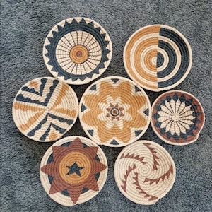 Puede incluir: Siete piezas redondas de decoraci&oacute;n de pared tejidas con diferentes patrones geom&eacute;tricos en negro, marr&oacute;n y beige.