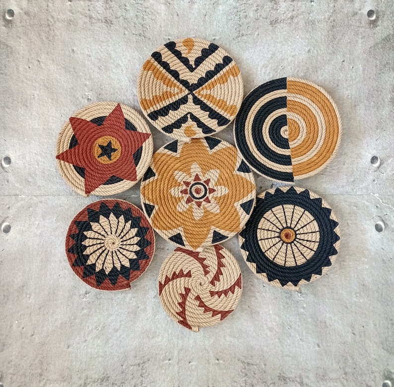 Puede incluir: Conjunto de siete piezas de decoraci&oacute;n de pared tejidas en varios patrones geom&eacute;tricos. Las piezas est&aacute;n hechas de materiales naturales y presentan una combinaci&oacute;n de colores negro, marr&oacute;n y beige.