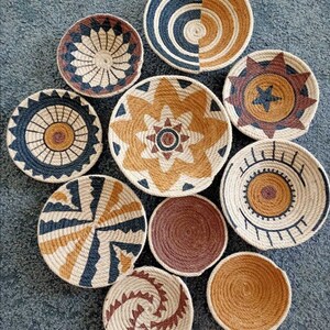 Puede incluir: Conjunto de nueve platos de pared tejidos con varios patrones geom&eacute;tricos. Los platos est&aacute;n hechos de materiales naturales y presentan una combinaci&oacute;n de colores marr&oacute;n, tostado y negro.