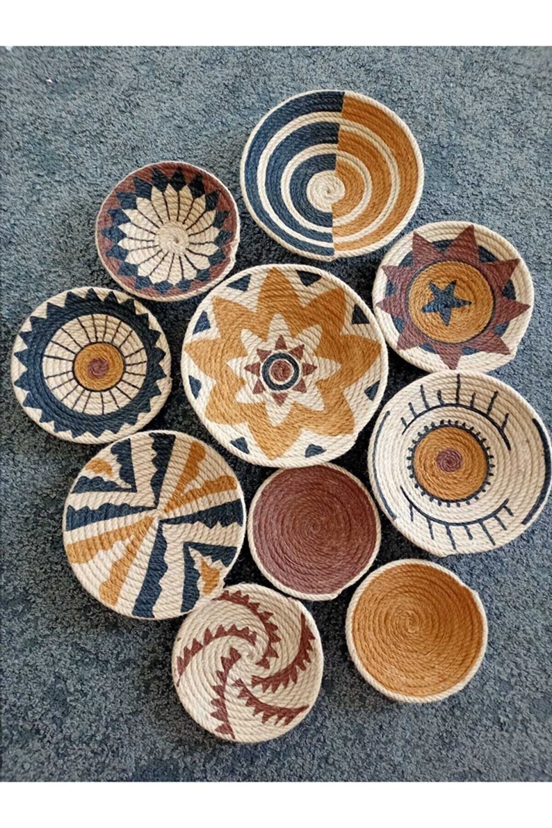 Puede incluir: Conjunto de nueve platos de pared tejidos con varios patrones geom&eacute;tricos. Los platos est&aacute;n hechos de materiales naturales y presentan una combinaci&oacute;n de colores marr&oacute;n, tostado y negro.