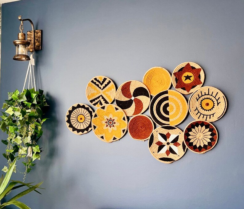 Puede incluir: Una colecci&oacute;n de 10 piezas redondas de decoraci&oacute;n de pared tejidas en varios patrones y colores, incluyendo negro, blanco, amarillo y marr&oacute;n. Las piezas est&aacute;n dispuestas en forma de nube en una pared azul.