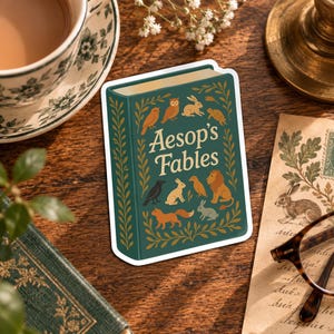 May include: A rectangular sticker featuring a dark green book titled Aesop's Fables in cream lettering. The cover is decorated with illustrations of an owl, rabbit, tortoise, crow, fox, and lion, all framed by a golden leaf border design.