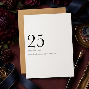 May include: A white greeting card with black text that reads 25, followed by the phonetic pronunciation [twen.ti'faiv] noun, and the phrase you're finally old enough to rent a car. The card is placed against a tan envelope on a dark surface.