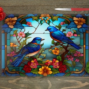A square glass cutting board with a stained-glass style image of two bluebirds on flowering branches, surrounded by a red bell pepper, a green bell pepper, and a clove of garlic, with a red-handled knife at the top right, all on a wooden surface.