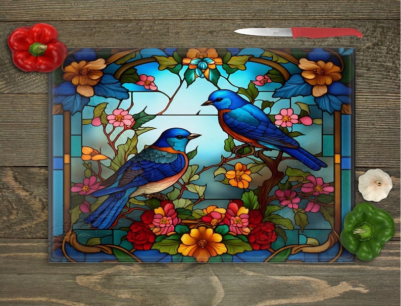 A square glass cutting board with a stained-glass style image of two bluebirds on flowering branches, surrounded by a red bell pepper, a green bell pepper, and a clove of garlic, with a red-handled knife at the top right, all on a wooden surface.